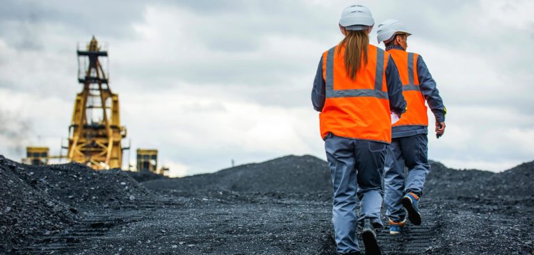 Two mining workers wearing high-vis jackets walking across the surface of an underground mine with mining machinery blurred in the distance