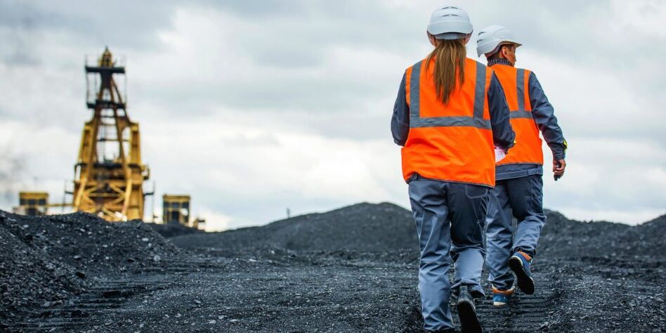 Two mining workers wearing high-vis jackets walking across the surface of an underground mine with mining machinery blurred in the distance